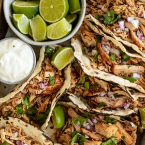An overview shot of instant pot carnitas in tacos next to onions, limes, and sour cream on a wood backrgound