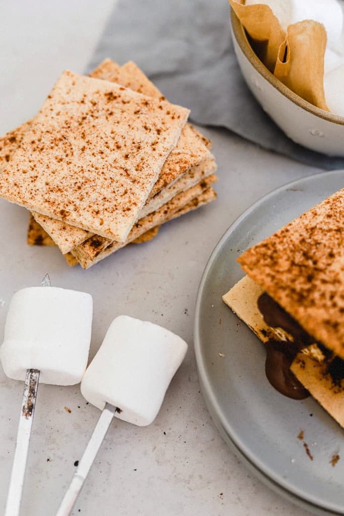 A s'more on a grey plate next to a stack of graham crackers and marshmallows on roasting sticks