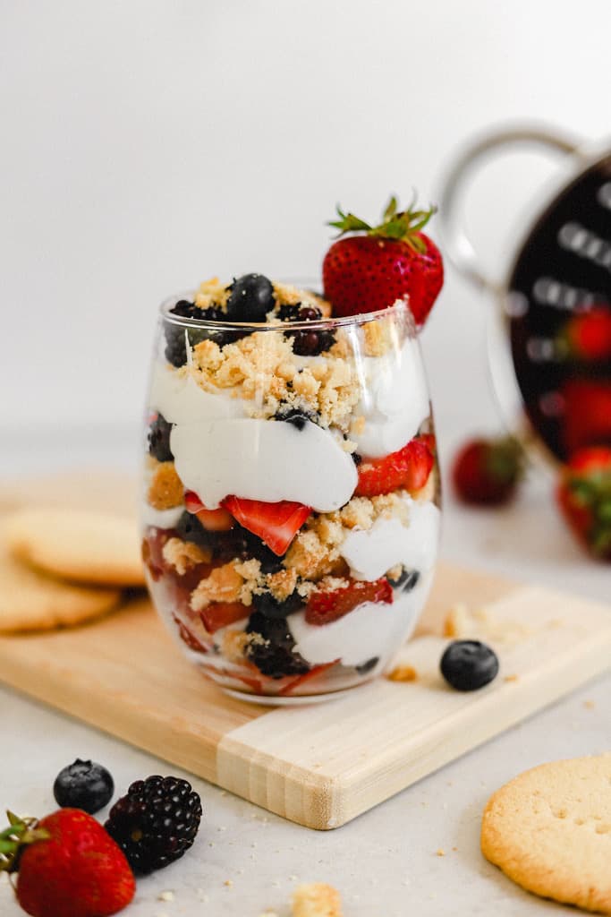 A glass with layers of berries, whipped coconut cream, and paleo shortbread cookie crumbles on a cutting board next to cookies and berries.