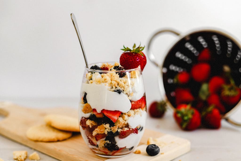 A glass with layers of berries, whipped coconut cream, and paleo shortbread cookie crumbles on a cutting board next to cookies and berries.