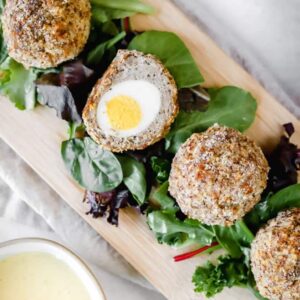 A top view of Scotch eggs with one cut open on top of greens and a cutting board next to a bowl of mustard sauce