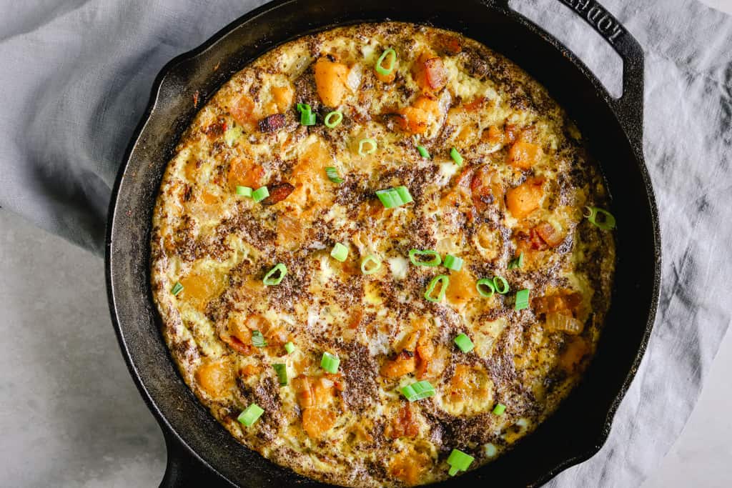 A cast iron pan with a whole30 kobacha squash frittata with bacon on a grey background