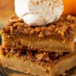 A stack of pumpkin pecan pie bars on a black plate topped with whipped cream and pumpkin pie spice