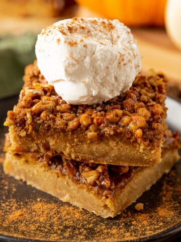 A stack of pumpkin pecan pie bars on a black plate topped with whipped cream and pumpkin pie spice