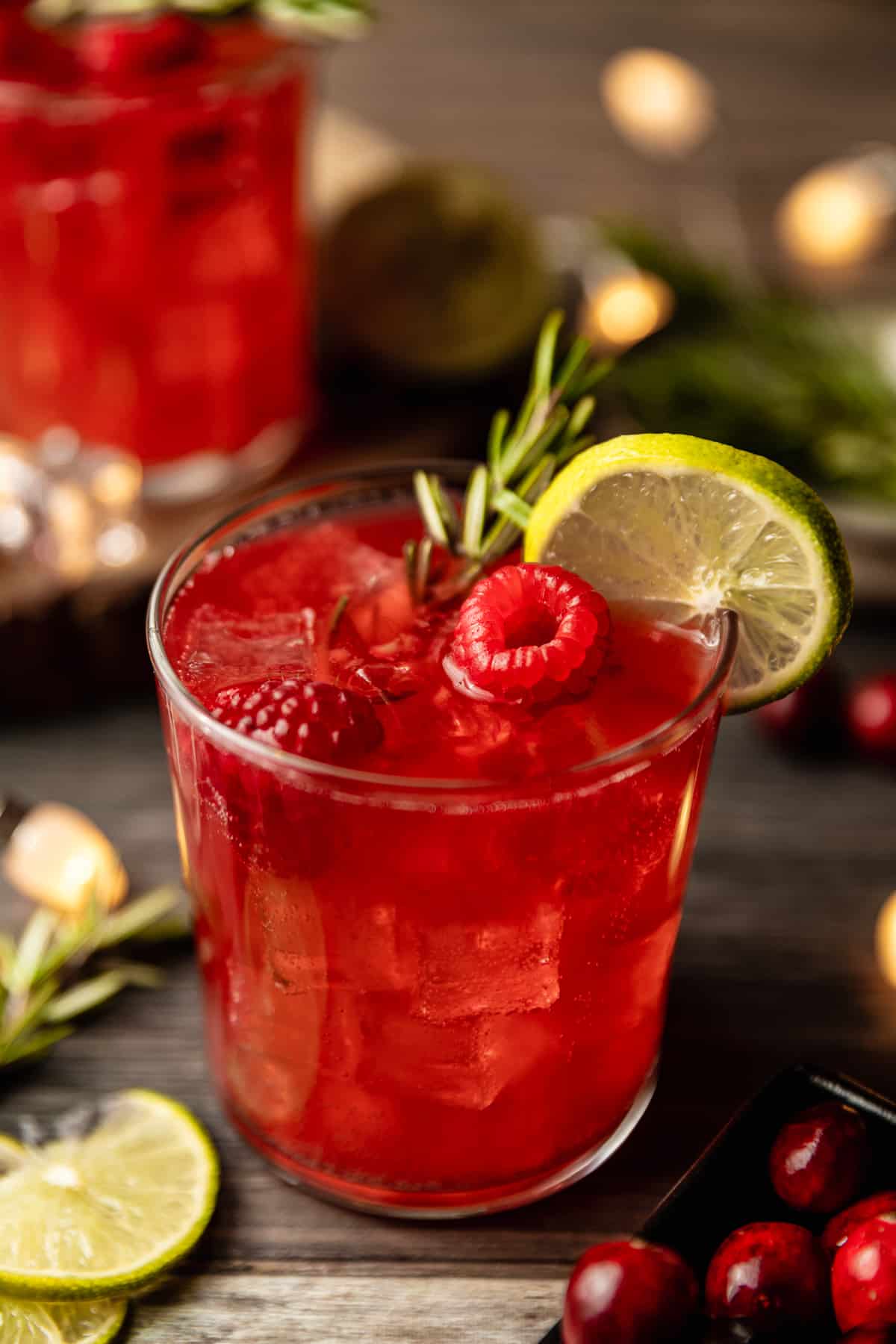 A glass filled with a cranberry raspberry mocktail topped with raspberries, rosemary, and limes on a wood background