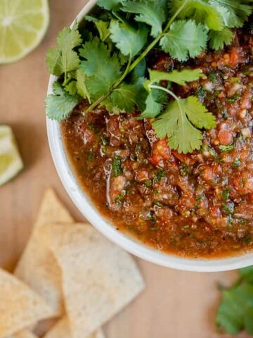 A bowl of salsa on a cutting board with chips, cilantro, jalapenos, and onion