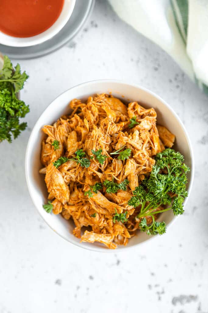 An overview shot of a bowl of buffalo chicken on a grey background next to greens and buffalo sauce