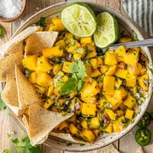 A top down shot of a bowl of mango salsa topped with cilantro, lime, and served with chips on a wood background.