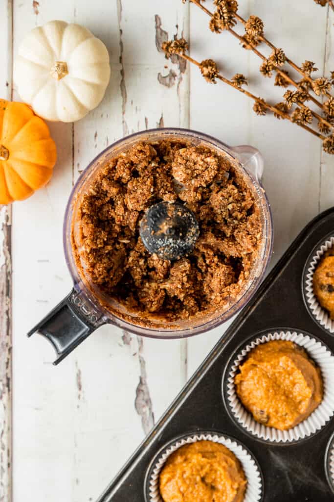 A mini food processor containing the ingredients for a paleo crumb topping near a muffin tin full of paleo pumpkin muffin batter