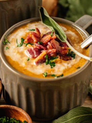 A bowl of Butternut Squash Soup topped with coconut cream, bacon, and fresh sage on a wood background