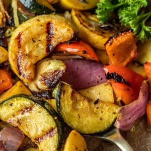 Smoked vegetables with grill marks being scooped by a gold spoon near parsley and peppercorns.