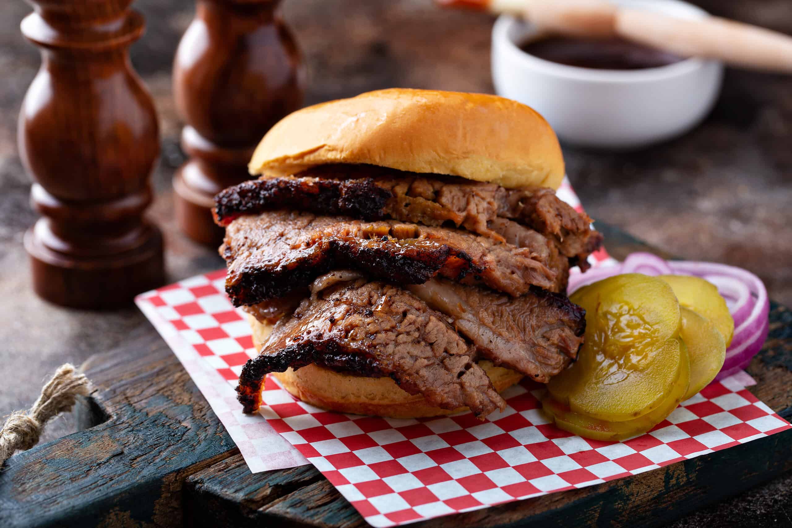 A sliced brisket sandwhich on a red and white checkered background next to bbq sauce.