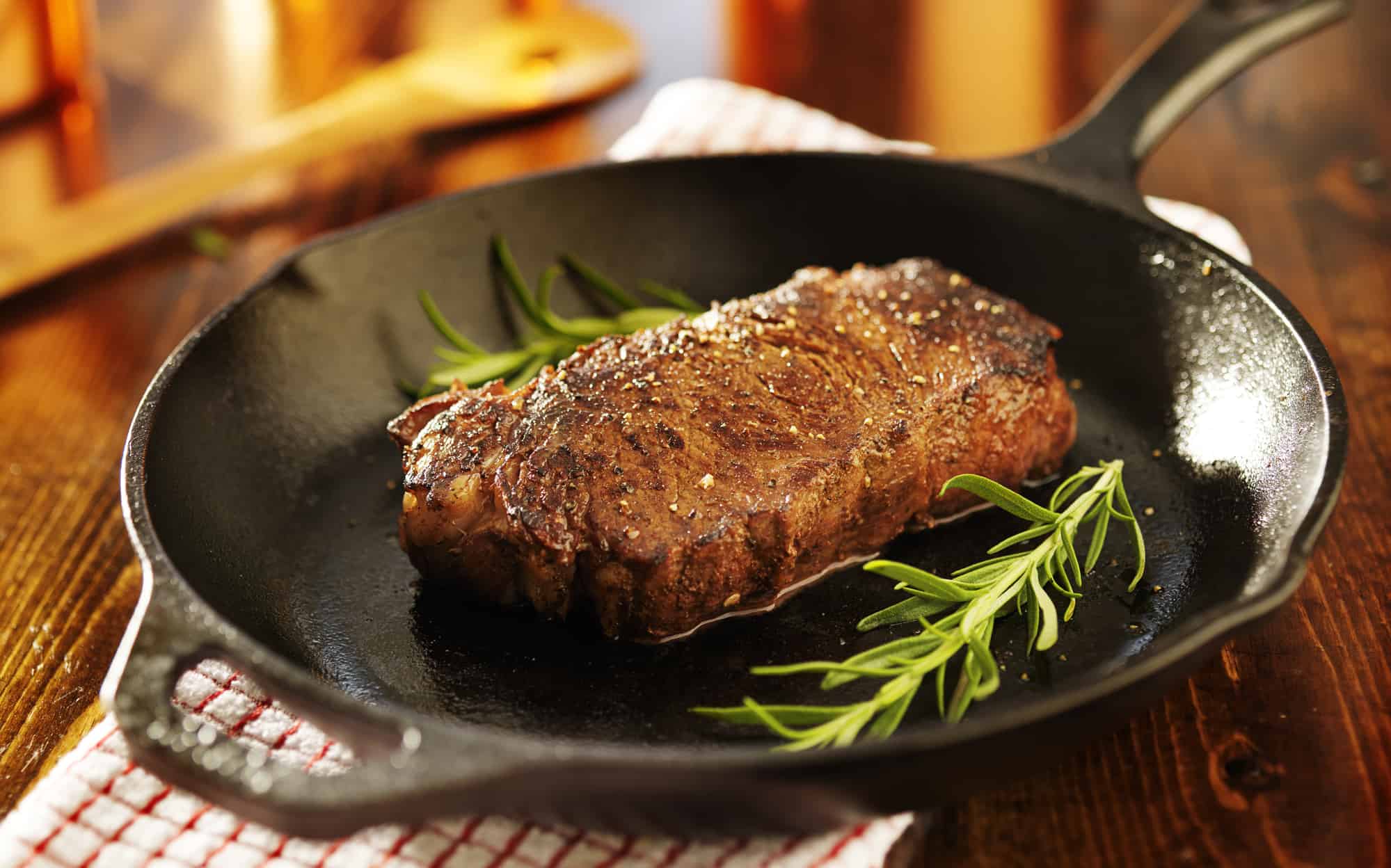 A cooked new york strip steak in a cast-iron skillet on a wood background topped with rosemary.