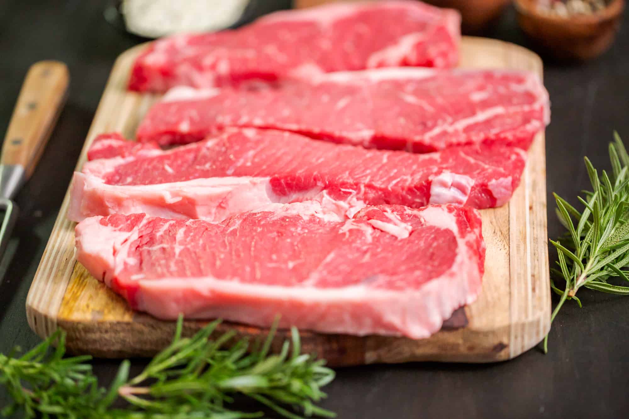 Raw NY Strip Steaks on a wood background near rosemary.